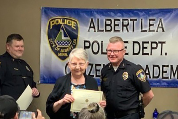 An elderly woman receives a certificate from a police officer at an event, standing in front of an 