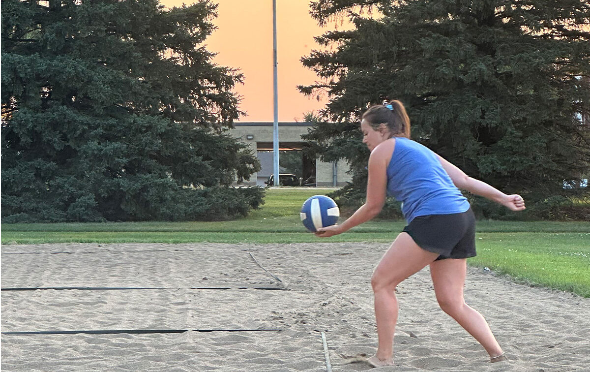 A woman in athletic clothing prepares to serve a volleyball on an outdoor sand court with trees and a building in the background.