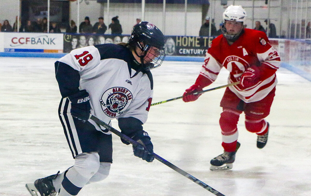 Two ice hockey players, one in a white and black uniform and the other in red and white, compete for the puck on an indoor rink.