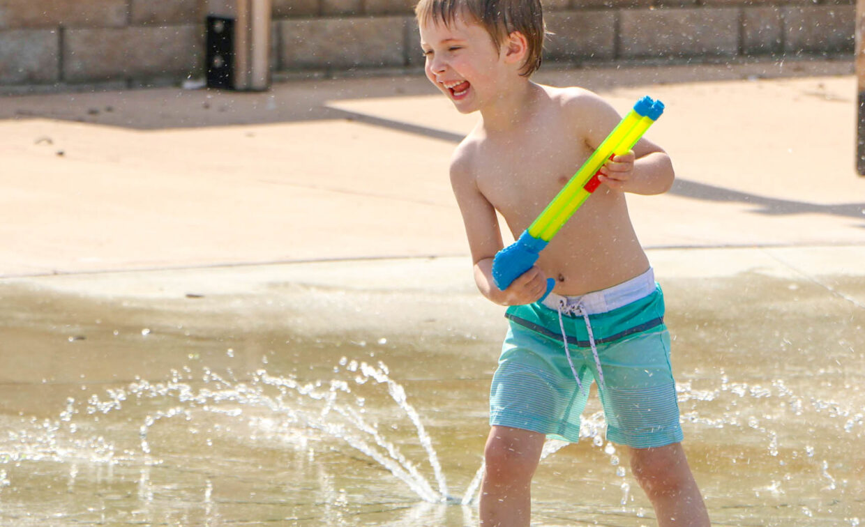 Young boy in swim trunks plays with a water gun in a splash pad area, surrounded by water sprays, on a sunny day.
