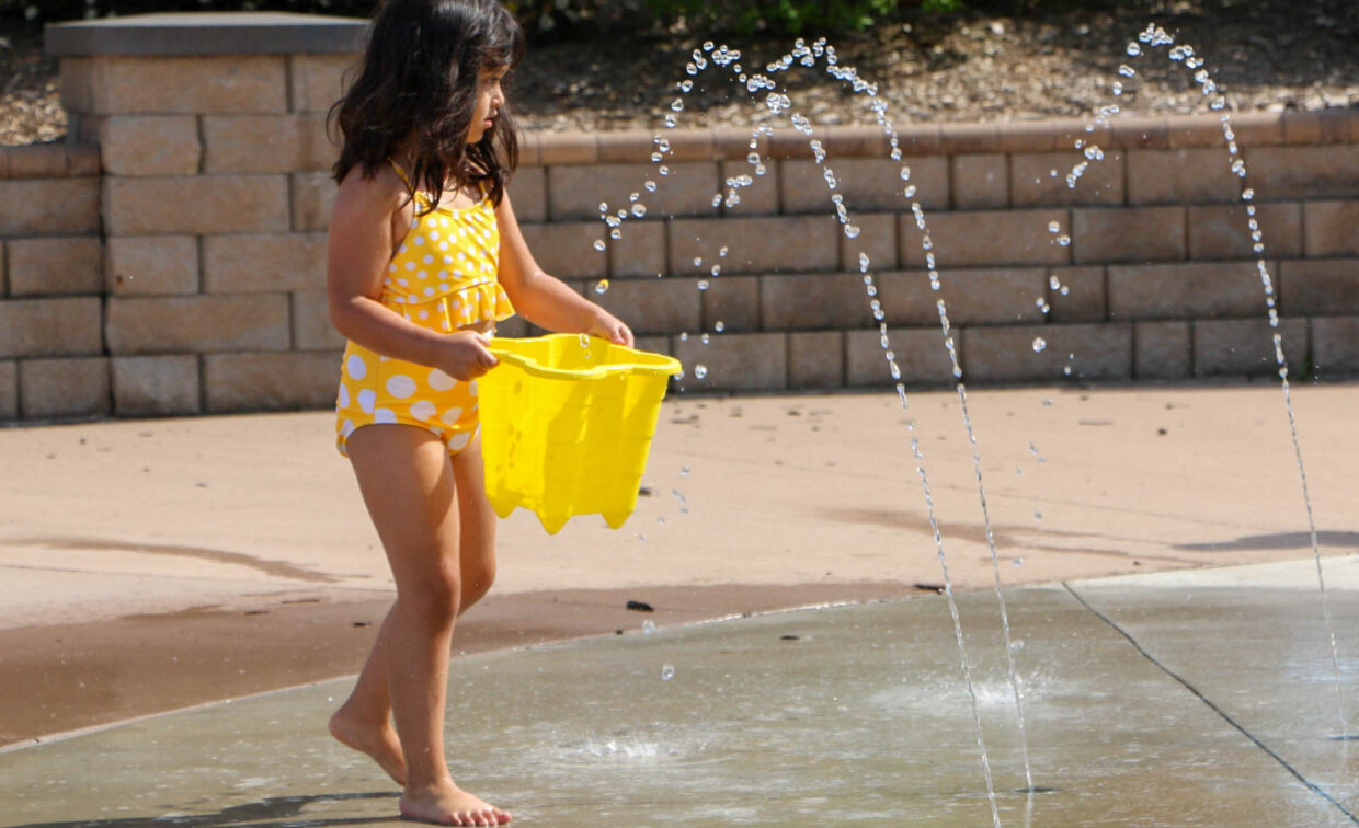 A young girl in a yellow polka dot swimsuit holds a yellow bucket while standing near water fountains at a splash pad.