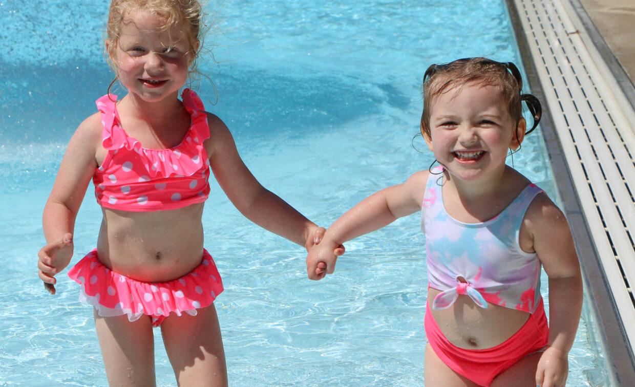 Two young children in swimsuits hold hands and smile while standing in shallow water at a swimming pool.