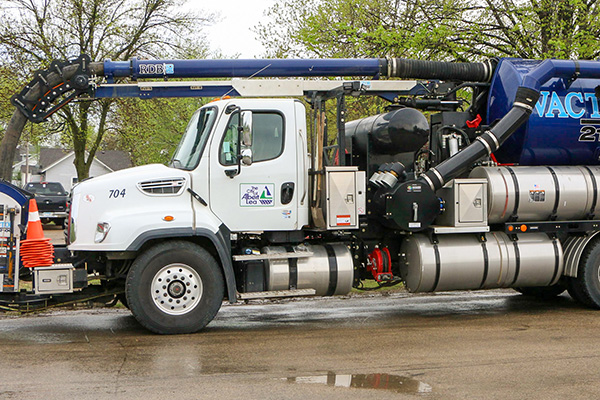 A vacuum truck is parked on a wet street with its vacuum hose extended and orange safety cones placed nearby.
