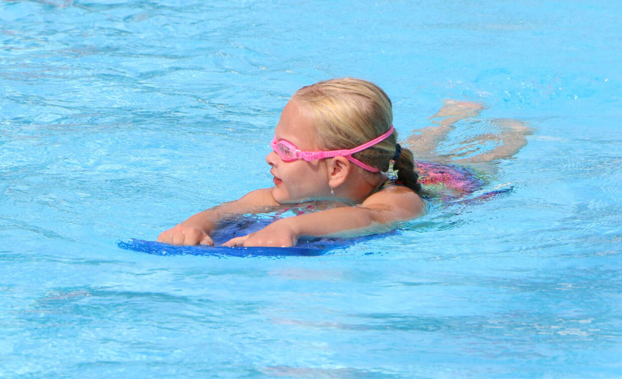 A young child wearing pink goggles swims in a pool using a blue kickboard.