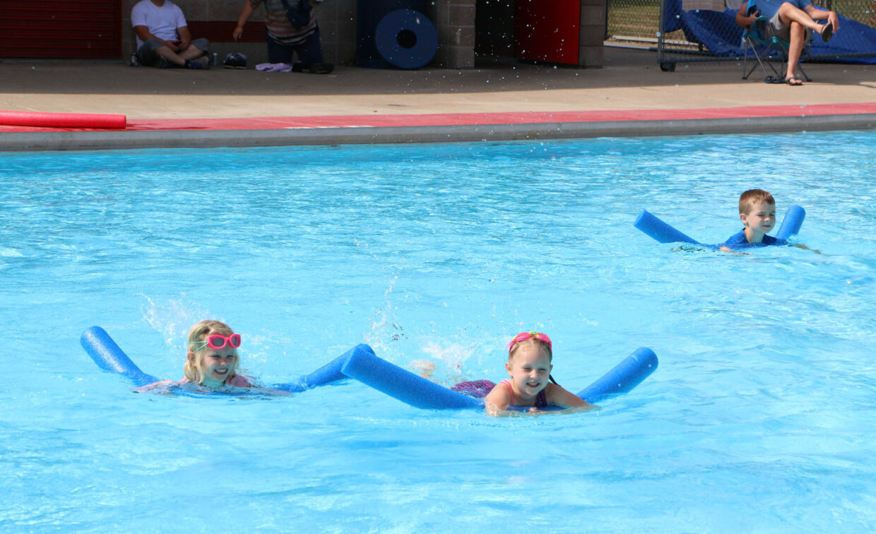 Three children use blue pool noodles to swim in an outdoor pool, with a few adults sitting in the background near the pool edge.