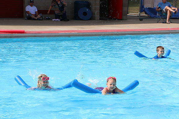 Three children use blue pool noodles to swim in an outdoor pool, with two adults watching from the poolside in the background.