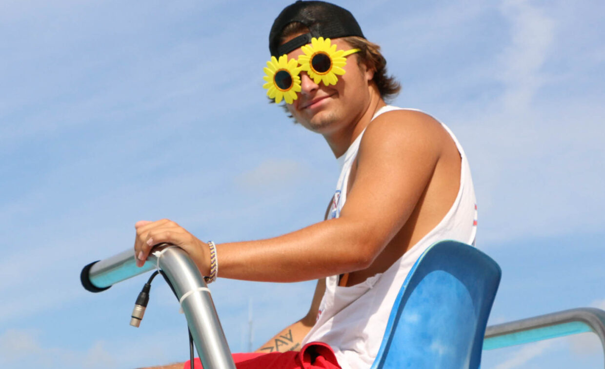 A person wearing sunflower-shaped sunglasses and a backward cap sits in a blue lifeguard chair under a partly cloudy sky.