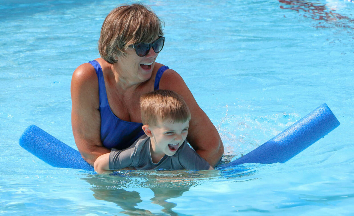 An older woman and a young boy use a blue pool noodle while swimming together in a pool. Both appear to be smiling.