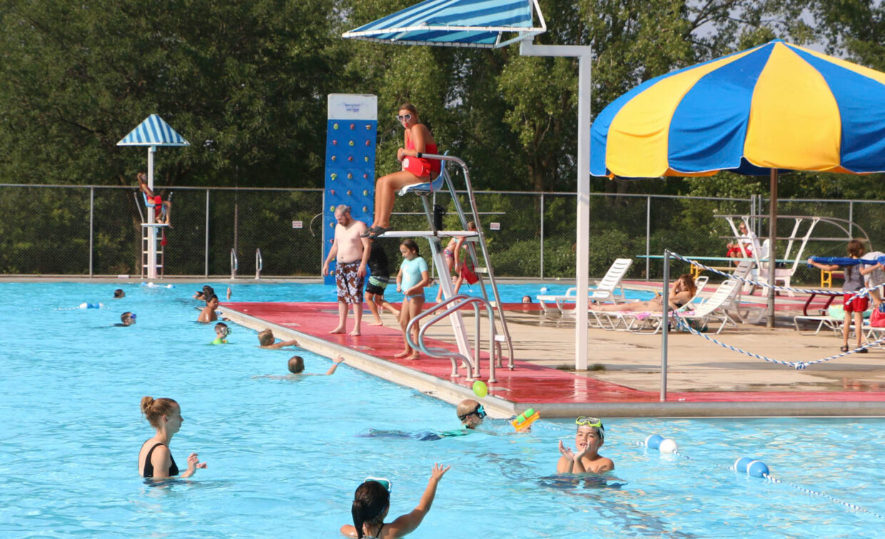 People are swimming and playing in a large outdoor pool with a lifeguard watching from a stand. Some people are seated under a yellow and blue umbrella near the poolside.