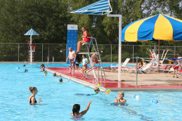 People are swimming and playing in a large outdoor pool with a lifeguard watching from a stand. Some people are seated under a yellow and blue umbrella near the poolside.