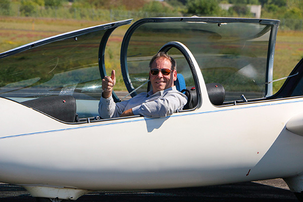 A man wearing sunglasses sits in the cockpit of a white glider, giving a thumbs-up gesture, with the canopy open and grassy background visible.