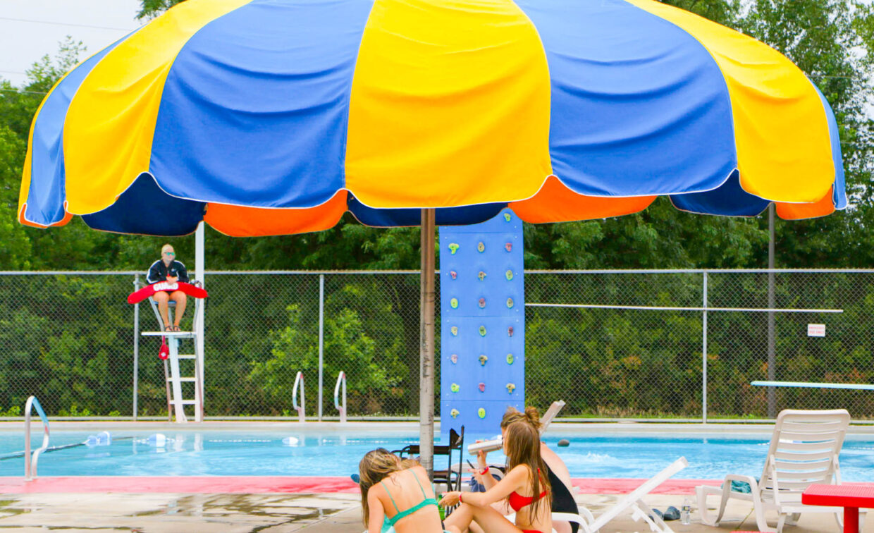 Three girls sit on lounge chairs under a large yellow and blue umbrella near a pool, with a lifeguard on duty and a climbing wall in the background.