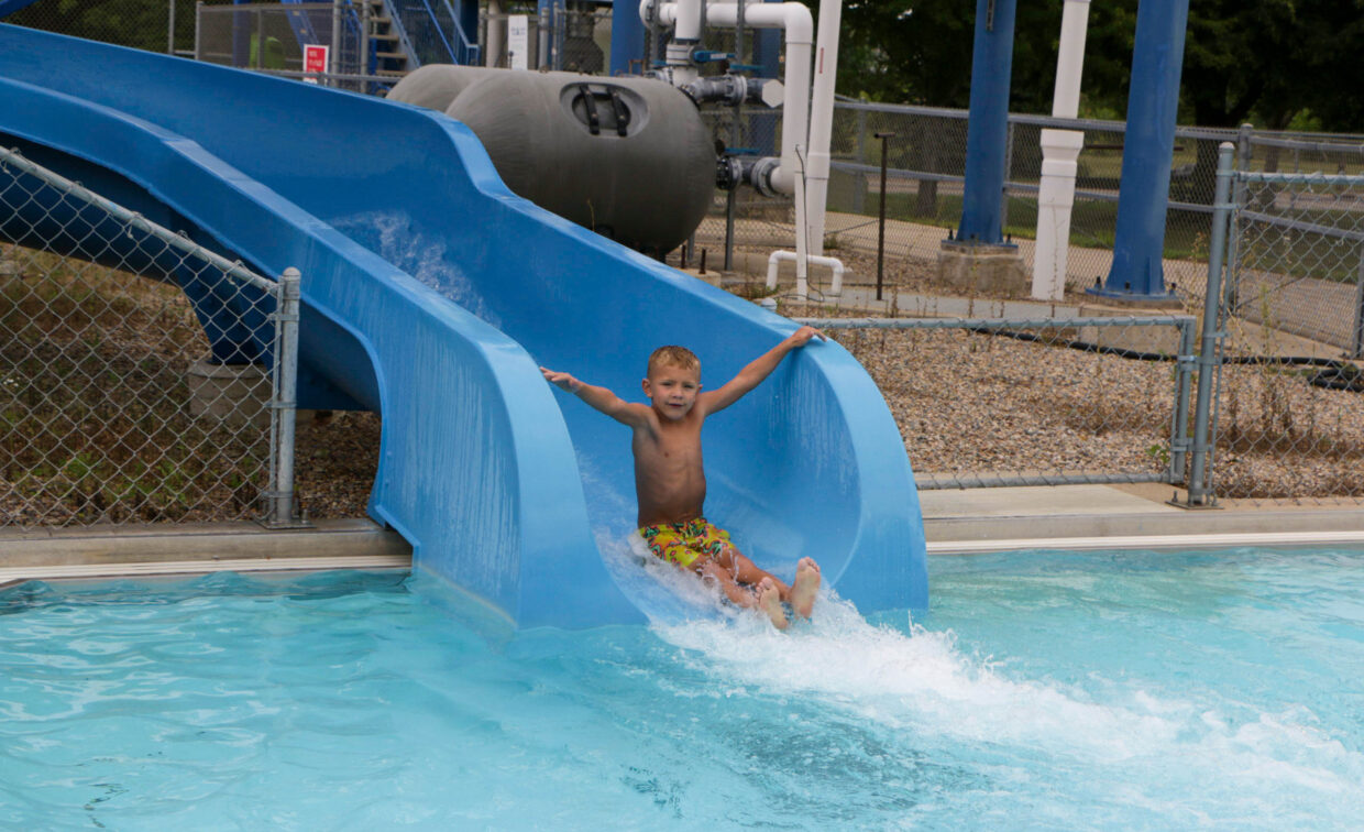 A young boy slides down a blue water slide into a pool, splashing water. Fence, pipes, and outdoor equipment are visible in the background.