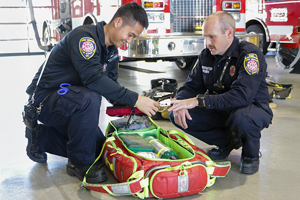 Two firefighters kneel beside an open emergency medical kit inside a fire station, preparing equipment in front of a fire truck.
