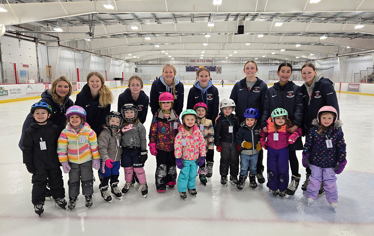 A group of children wearing ice skates and helmets pose with adults in jackets on an indoor ice skating rink.