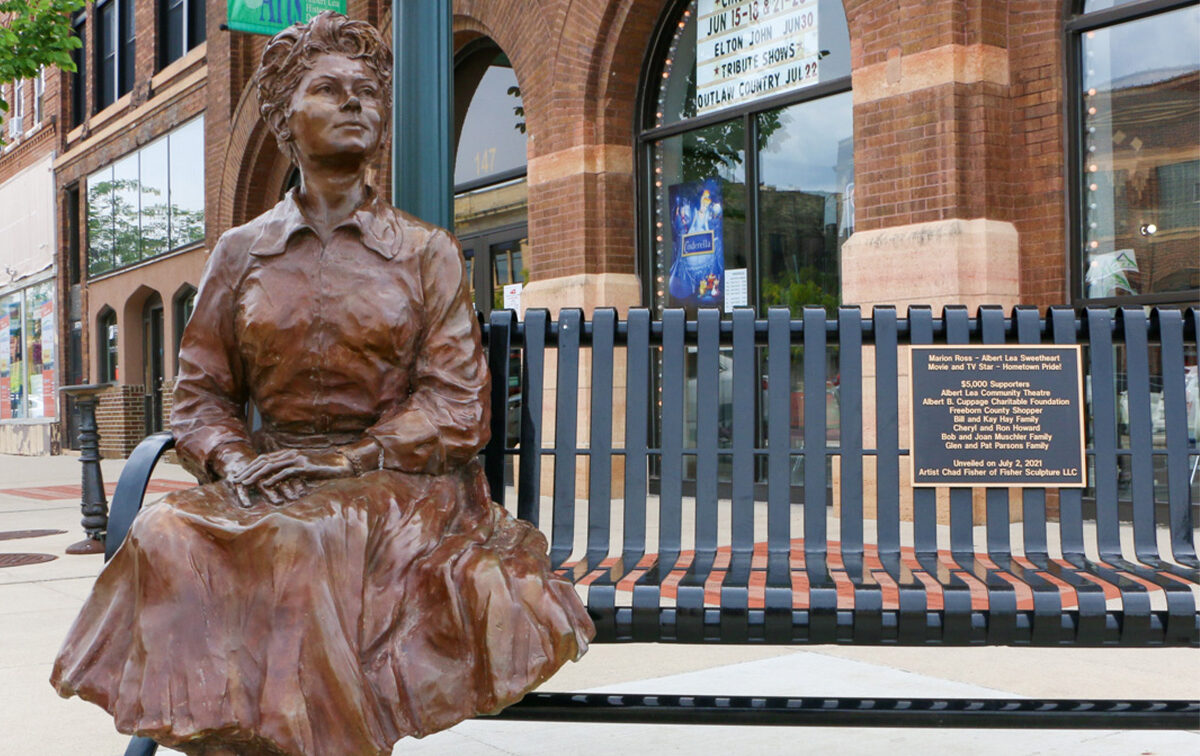 A bronze statue of a seated woman is positioned next to a metal bench with a commemorative plaque, outside a historic brick building with arched windows.