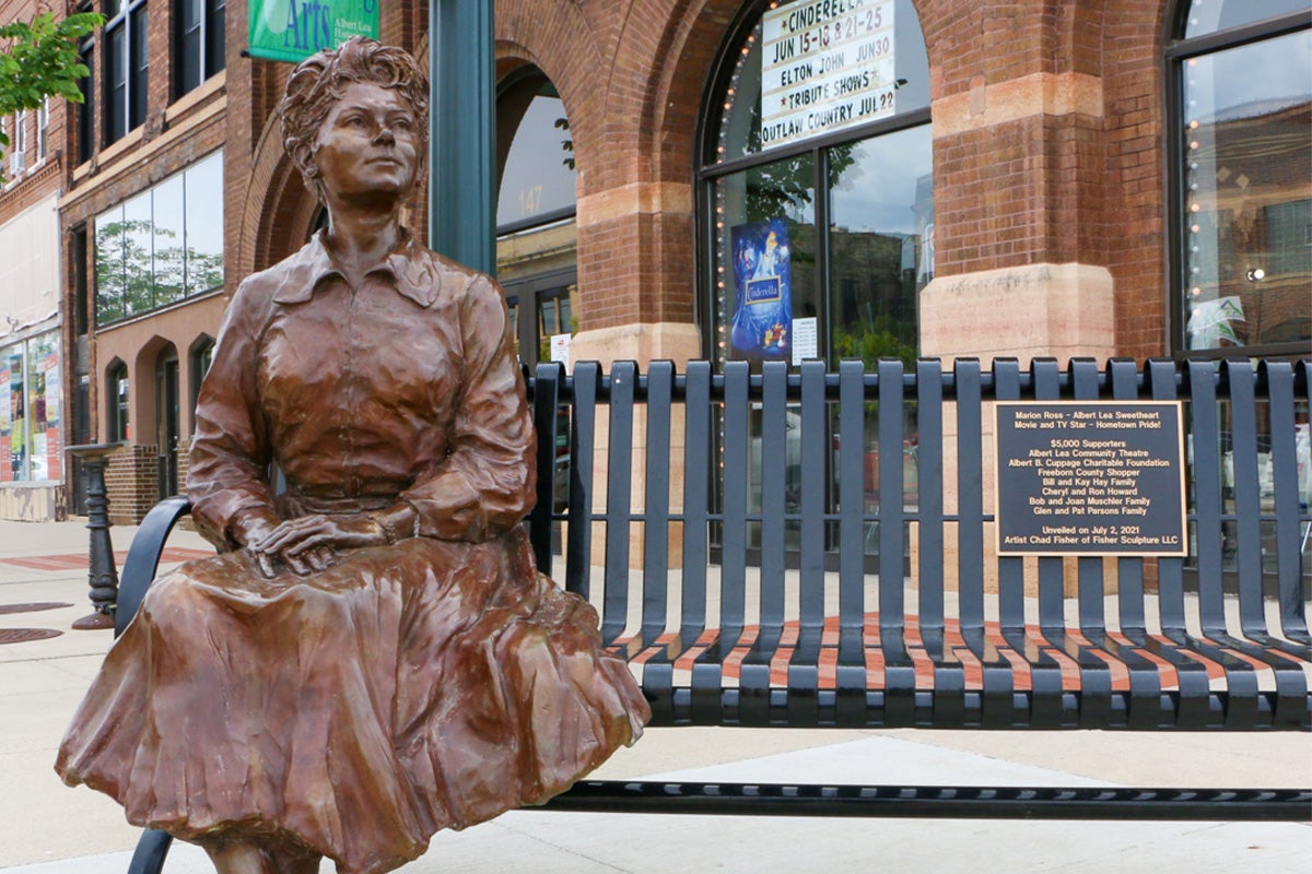 A bronze statue of a seated woman is positioned next to a metal bench with a commemorative plaque, outside a historic brick building with arched windows.
