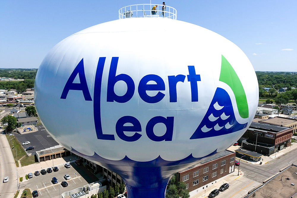 Large water tower with the words "Albert Lea" painted on it, overlooking buildings and streets in a small city.
