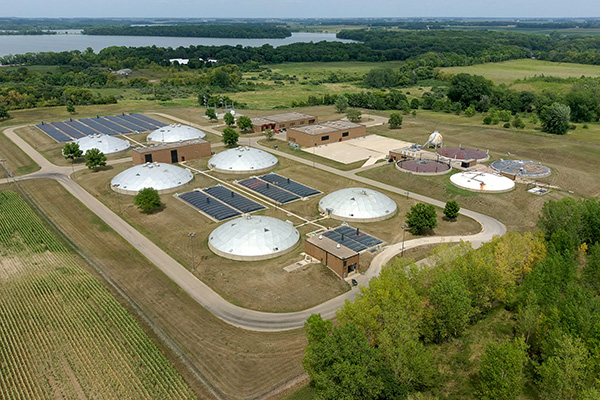 Aerial view of a wastewater treatment plant surrounded by fields and trees, featuring multiple circular and rectangular buildings with solar panels on some roofs.