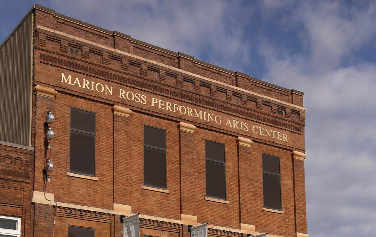 A brick building with the sign "Marion Ross Performing Arts Center" on the top facade, set against a partly cloudy sky.
