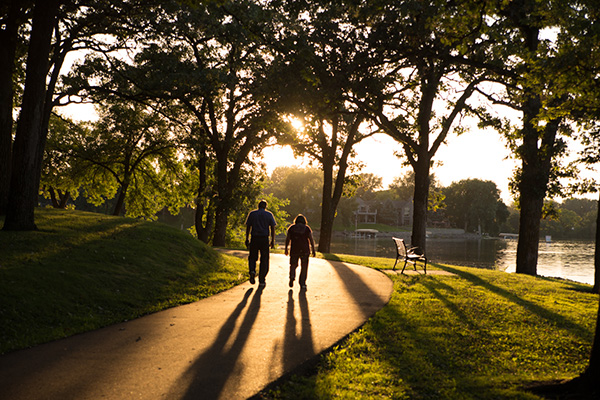 Two people walk on a tree-lined park path at sunset, casting long shadows. A bench and a river are visible in the background.