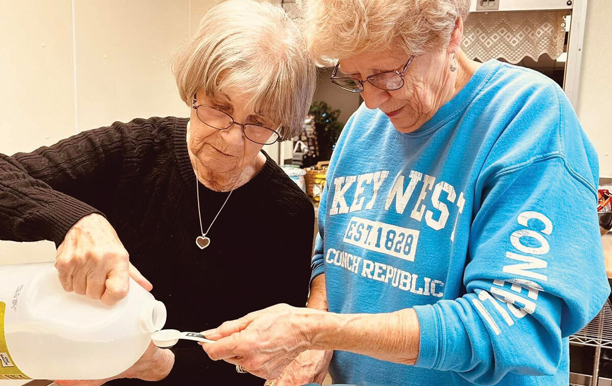 Two older women in a kitchen measure liquid from a jug into a spoon, working together on a cooking or baking task.