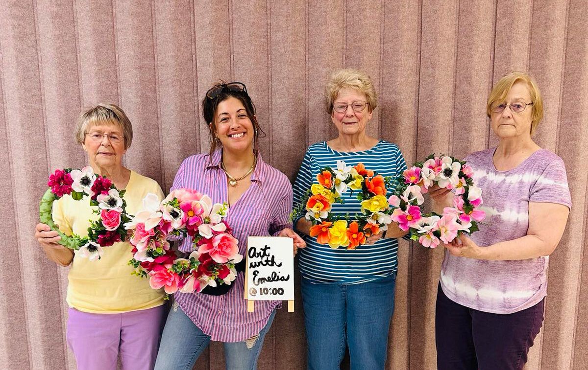 Four women stand in front of a pink curtain, each holding a colorful floral wreath. One woman holds a sign that says, "Art with Friends 10:00.