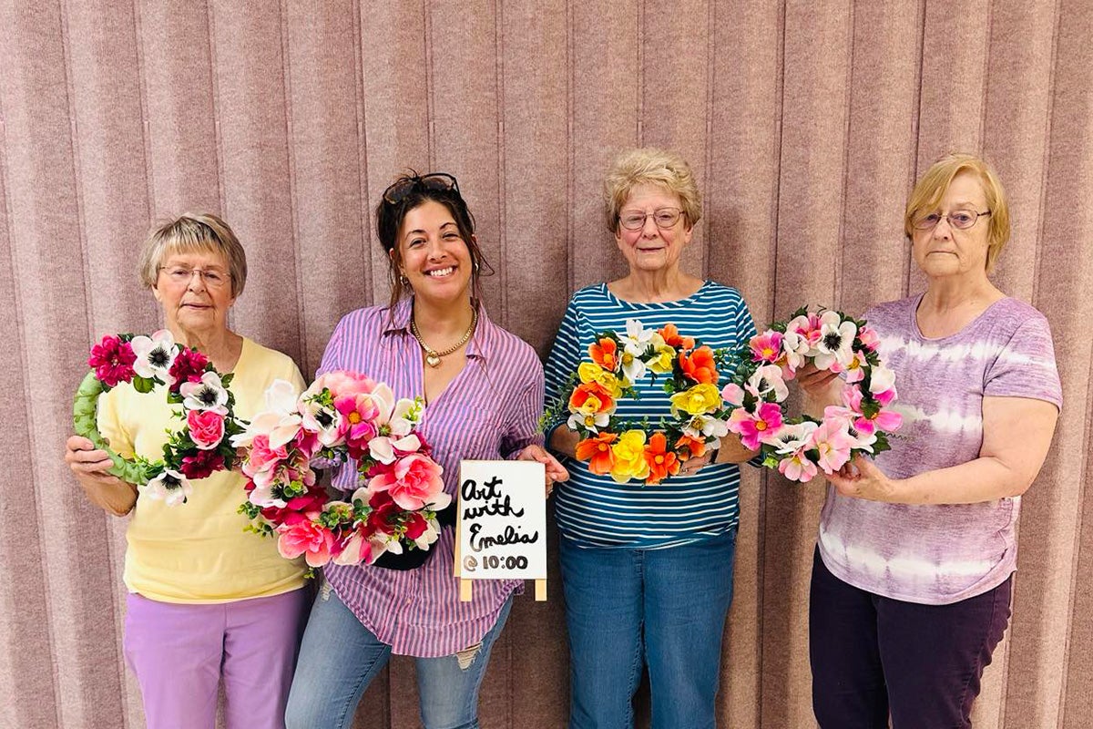 Four women stand in front of a pink curtain, each holding a colorful floral wreath. One woman holds a sign that says, 