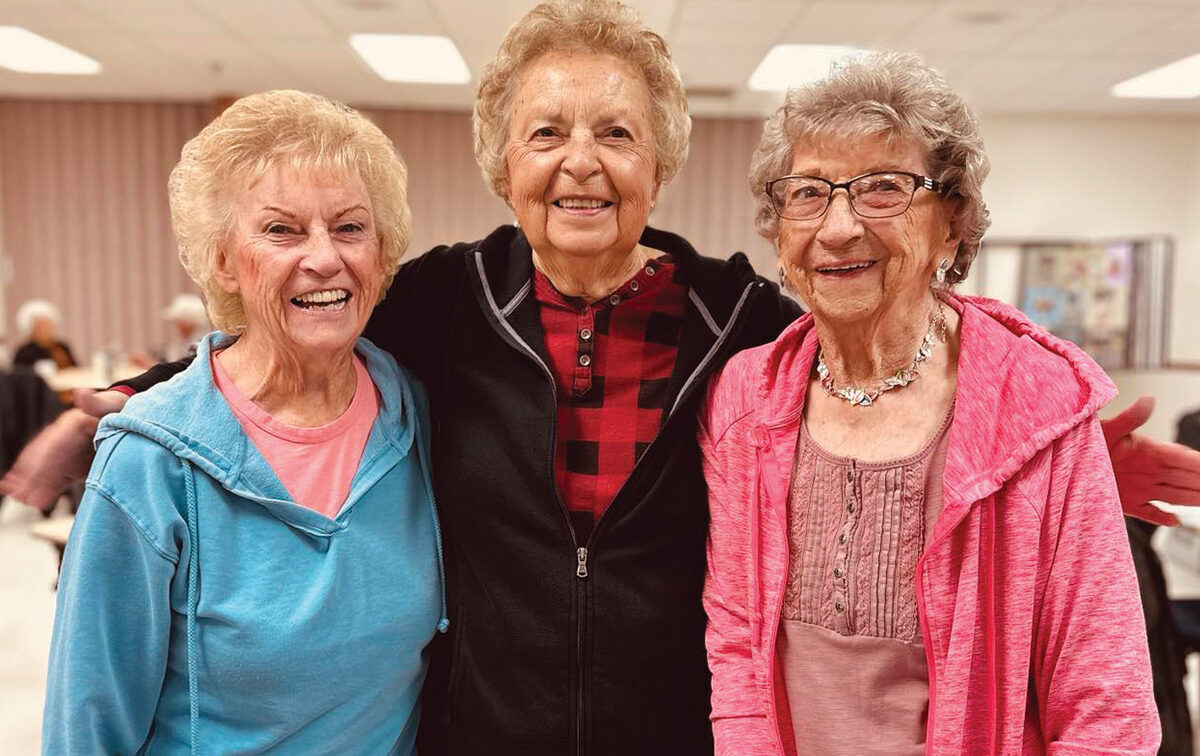 Three elderly women stand close together indoors, smiling at the camera. They are wearing casual clothing and glasses, and appear to be in a community or social gathering space.