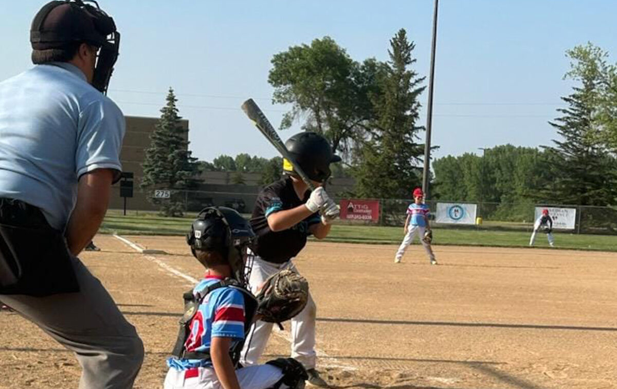 A youth baseball player prepares to bat as the catcher and umpire crouch behind home plate on an outdoor field. Other players are visible in the background.