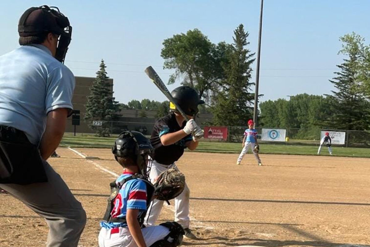 A youth baseball player prepares to bat as the catcher and umpire crouch behind home plate on an outdoor field. Other players are visible in the background.