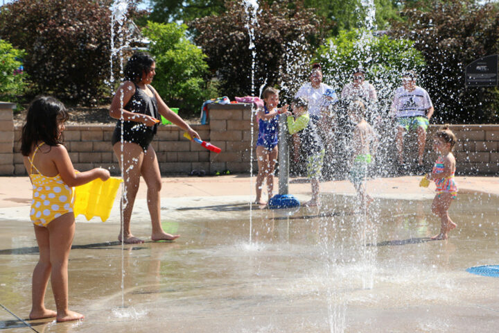 Children and an adult play in a water fountain area at a park on a sunny day, with others sitting on benches in the background.