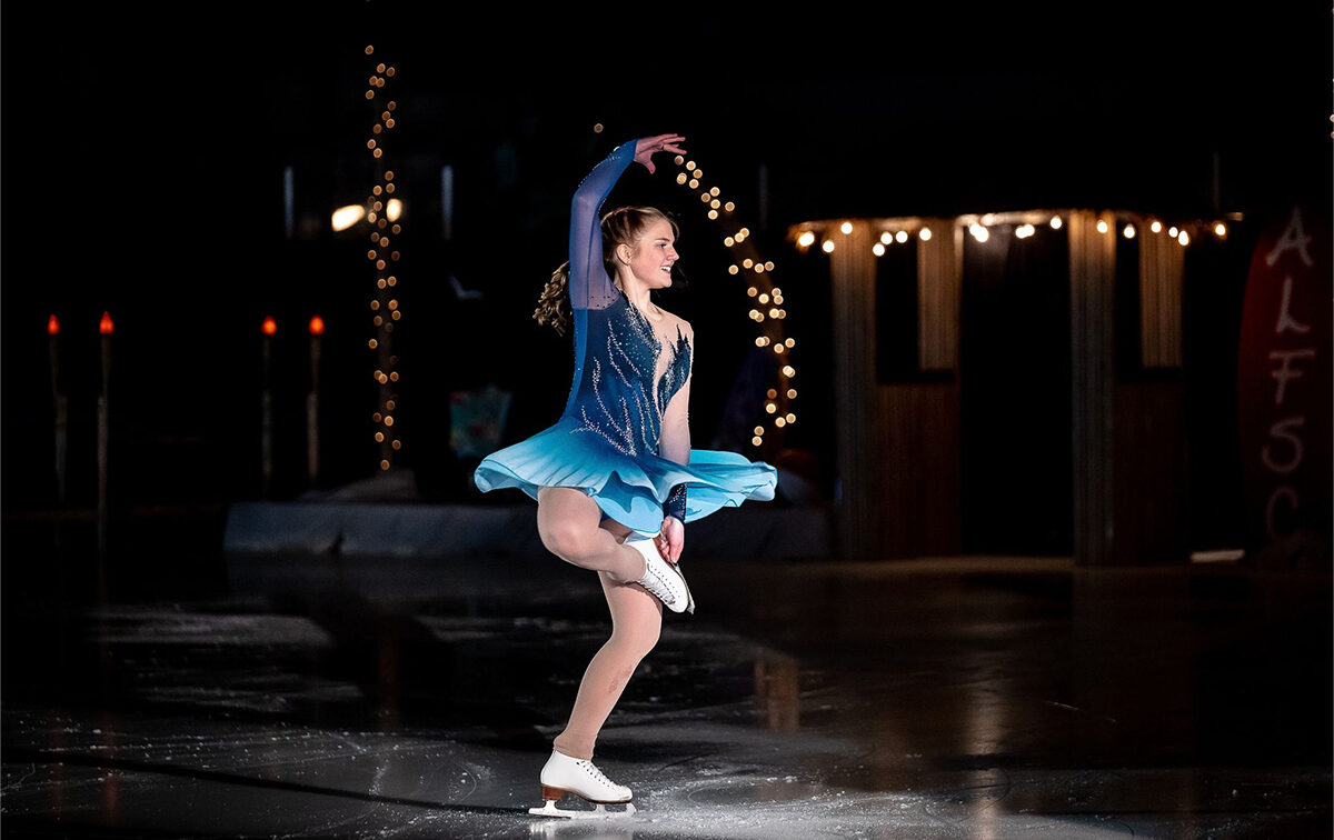 A figure skater in a blue costume performs a spin on an indoor ice rink, with string lights and dark background decorations visible.