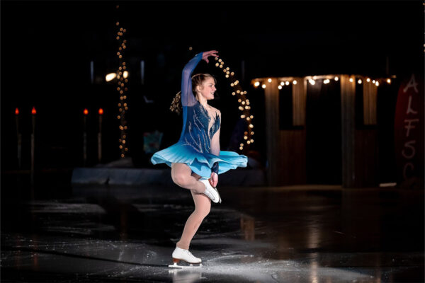 A figure skater in a blue costume performs a spin on an indoor ice rink, with string lights and dark background decorations visible.