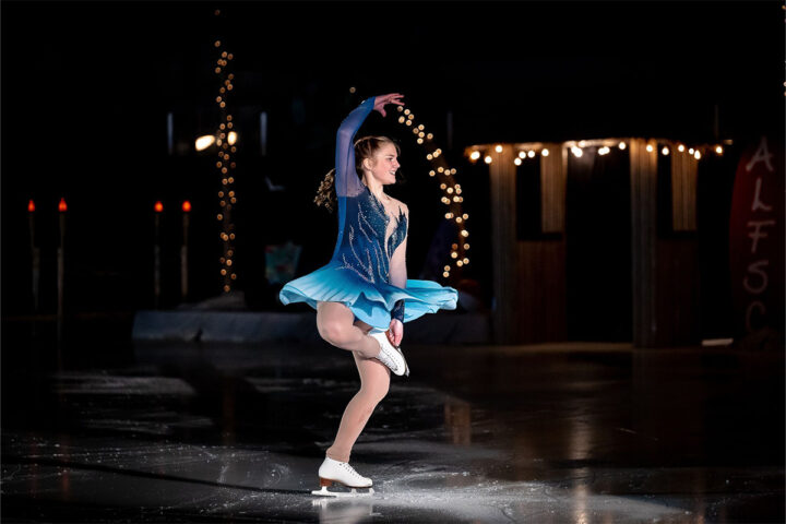 A figure skater in a blue costume performs a spin on an indoor ice rink, with string lights and dark background decorations visible.