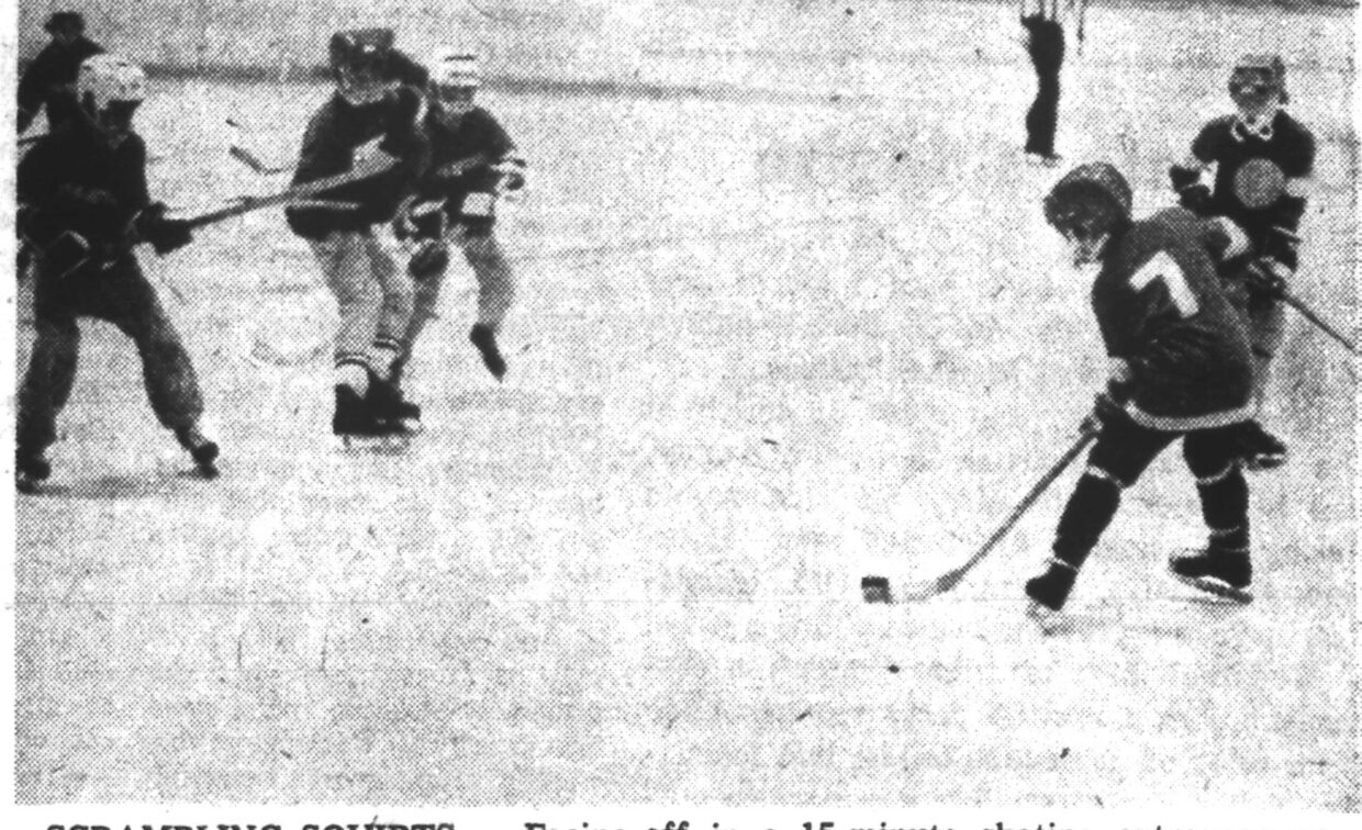 A group of young boys play an ice hockey game, skating and chasing the puck as a referee watches in the background.