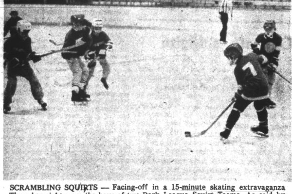 A group of young boys play an ice hockey game, skating and chasing the puck as a referee watches in the background.