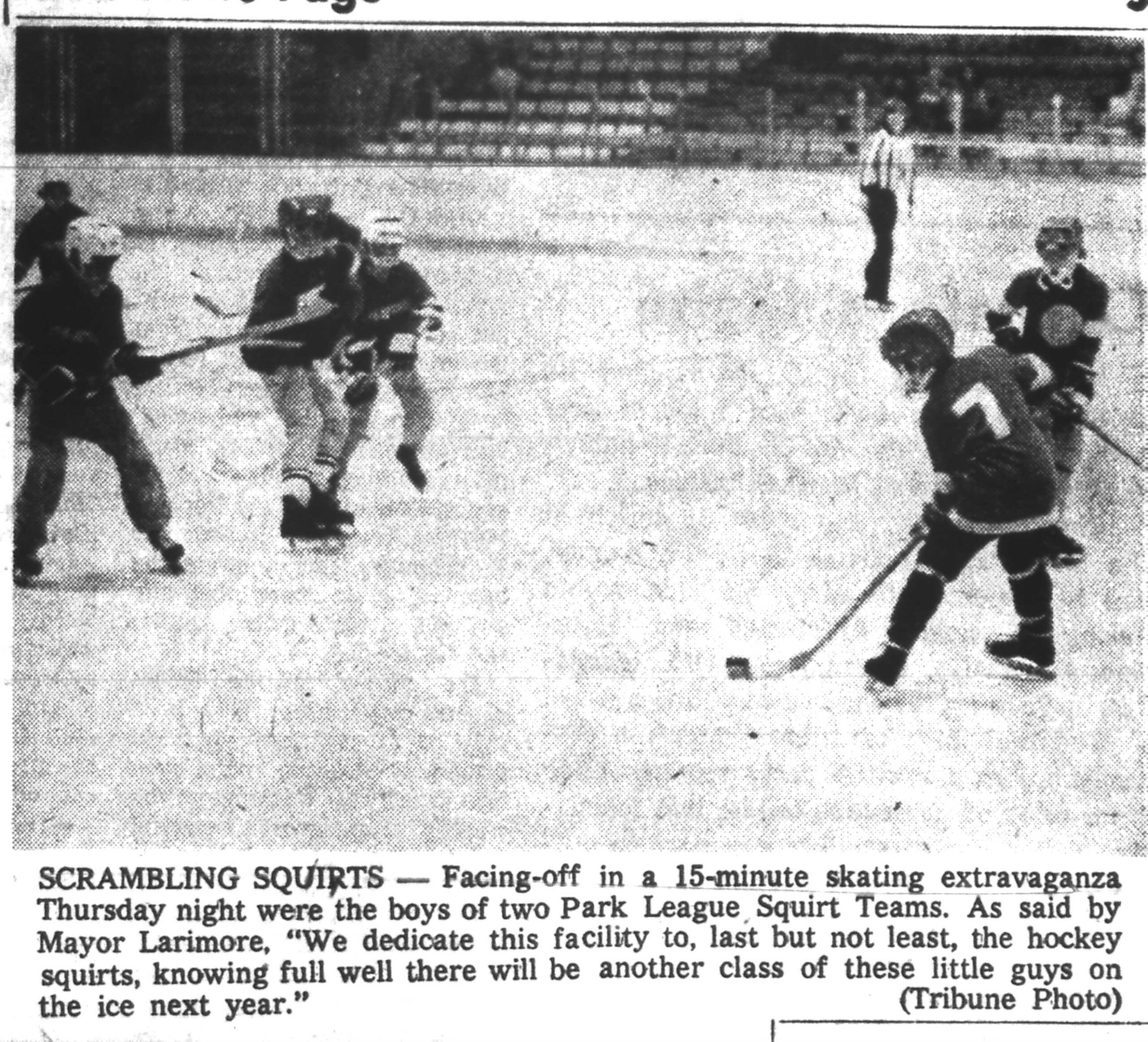 A group of young boys play an ice hockey game, skating and chasing the puck as a referee watches in the background.
