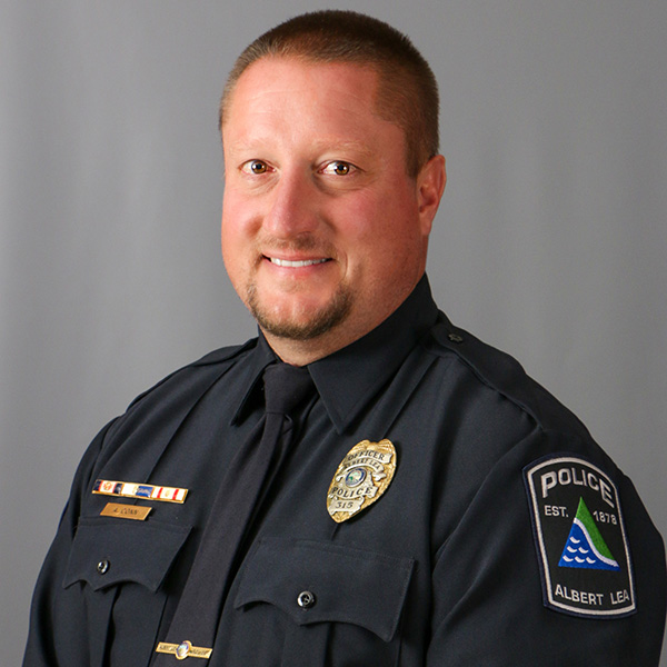 Adam Conn, A man in a police uniform with a badge and shoulder patch smiles, facing the camera against a plain background.