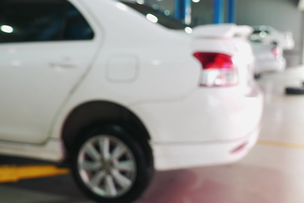 A blurred image shows the rear side of a white sedan parked indoors, with another white car visible in the background.