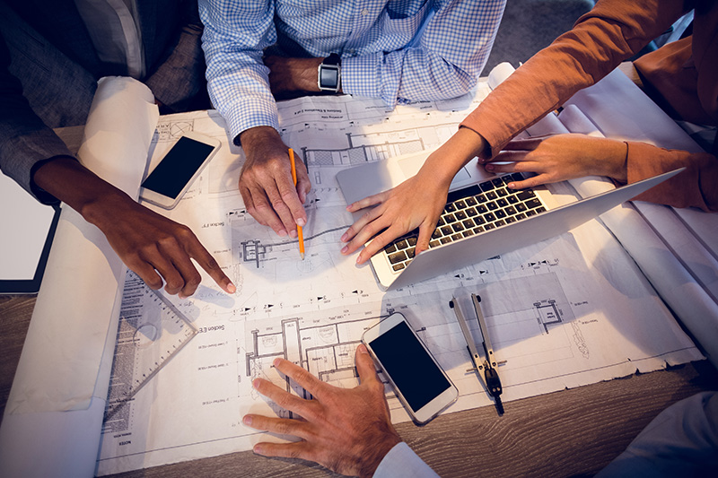 Four people work together on architectural blueprints at a table, with hands pointing, a laptop, smartphones, and drawing tools visible.