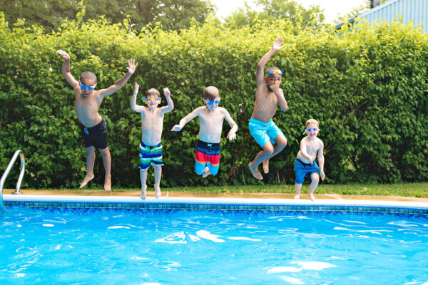 Five children in swimsuits and goggles jump into an outdoor swimming pool, mid-air, with greenery and a fence in the background.