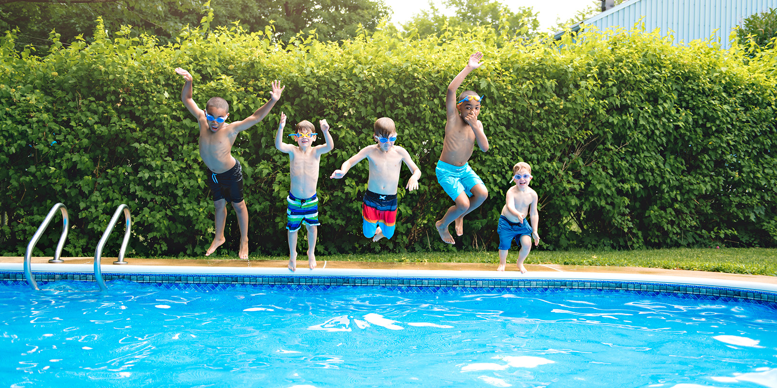 Five children in swimsuits and goggles jump into an outdoor swimming pool, mid-air, with greenery and a fence in the background.