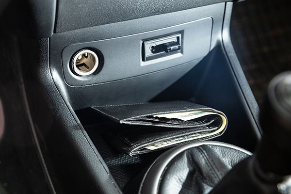 A black leather wallet with cash is placed in the center console compartment of a car, near the cigarette lighter and a switch.