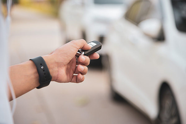 A person holding a car key remote is about to unlock a white car parked on the street.