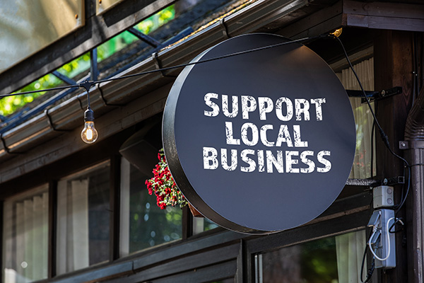 A round black sign with white text reading "Support Local Business" hangs outside a building near a lightbulb and a small bunch of red flowers.