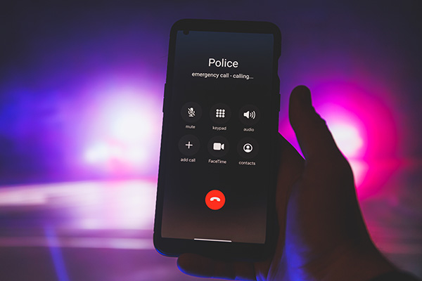 A hand holding a smartphone showing an active emergency call to the police, with blurred blue and pink lights in the background.