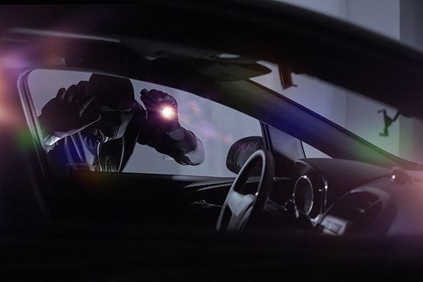 A person wearing a hoodie shines a flashlight while peering into the window of a parked car at night.