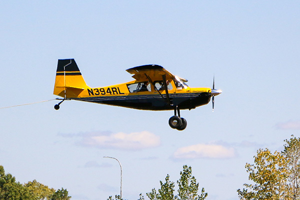 A yellow and black small aircraft is flying above the trees against a clear blue sky with a few clouds.