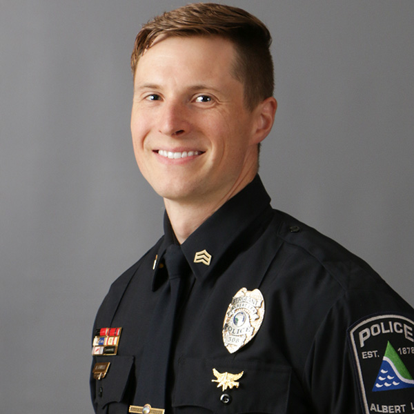 Alex Kunkel, Man in a police uniform with a smile, badges, and insignia on his shirt, standing against a gray background.